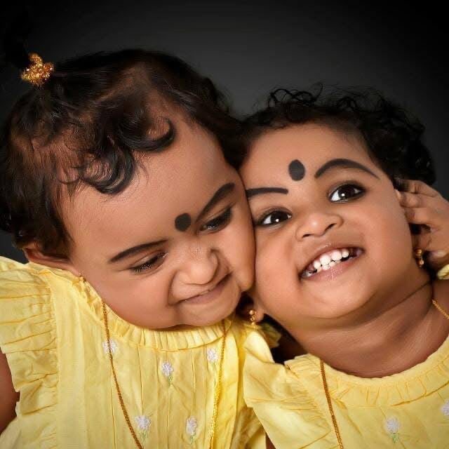 Studio portrait of two toddler sisters in matching yellow frocks with kajal and black bindis, cheek to cheek and laughing against a dark grey backdrop — kids photography Deira Dubai