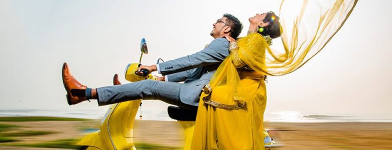 Couple riding a yellow vintage scooter along a coastal road, the woman in a yellow saree with her dupatta streaming in the wind — cinematic pre-wedding photography by Dar Al Muteena Studio