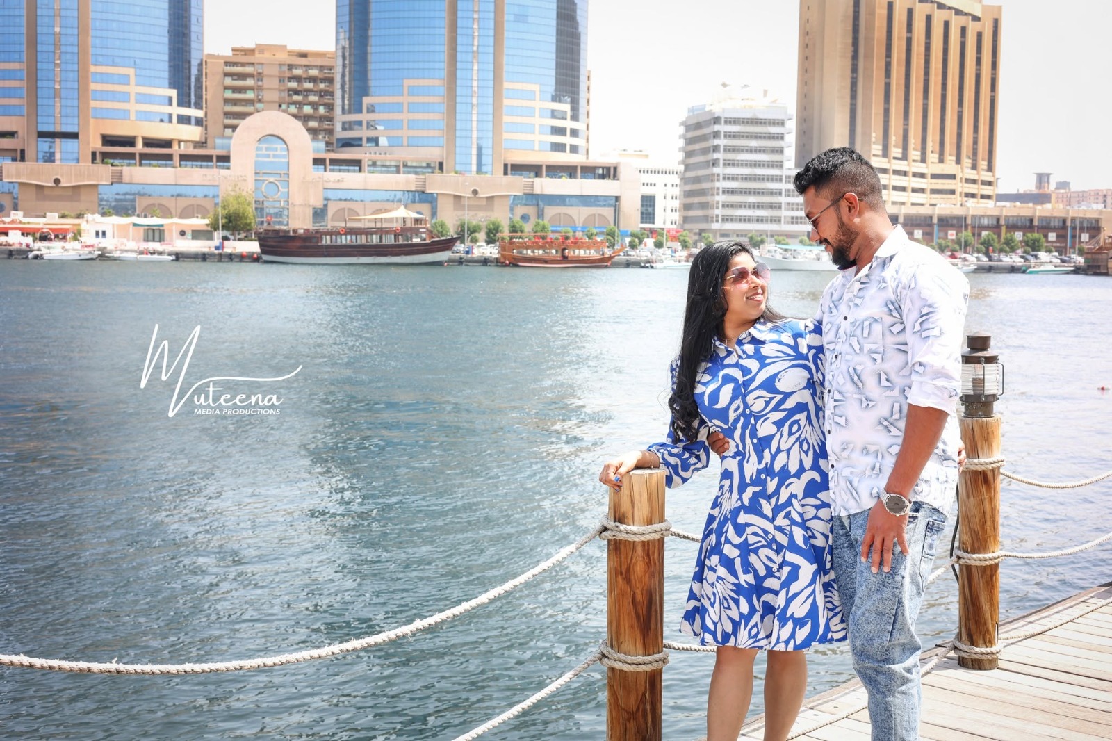 Pre-wedding couple portrait at Dubai Creek waterfront with a traditional wooden dhow and Deira skyscrapers behind, woman in a blue and white printed dress