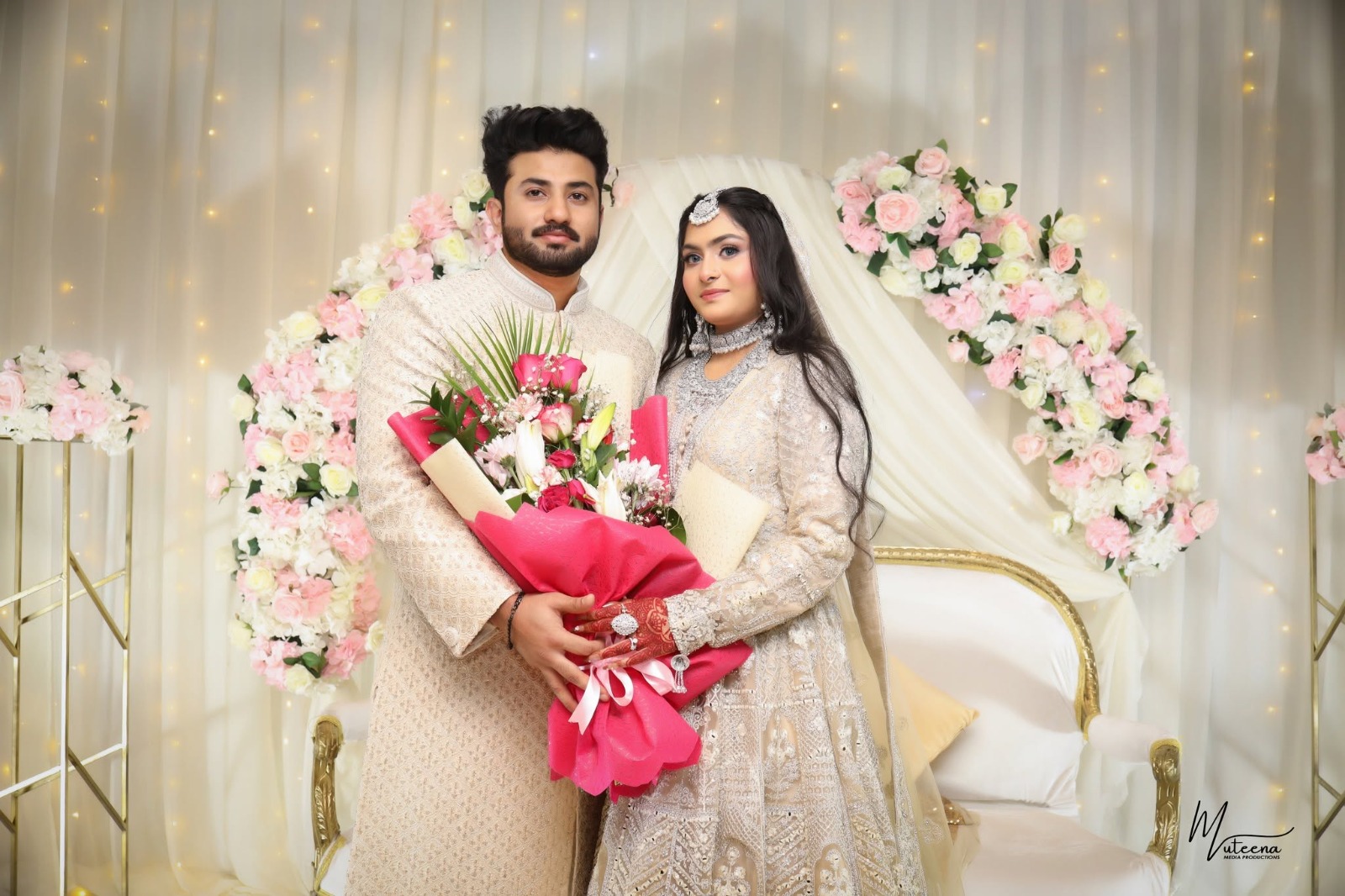 Full-length wedding portrait of a Muslim bride and groom on a floral nikkah stage in cream and ivory couture, holding a large pink rose bouquet