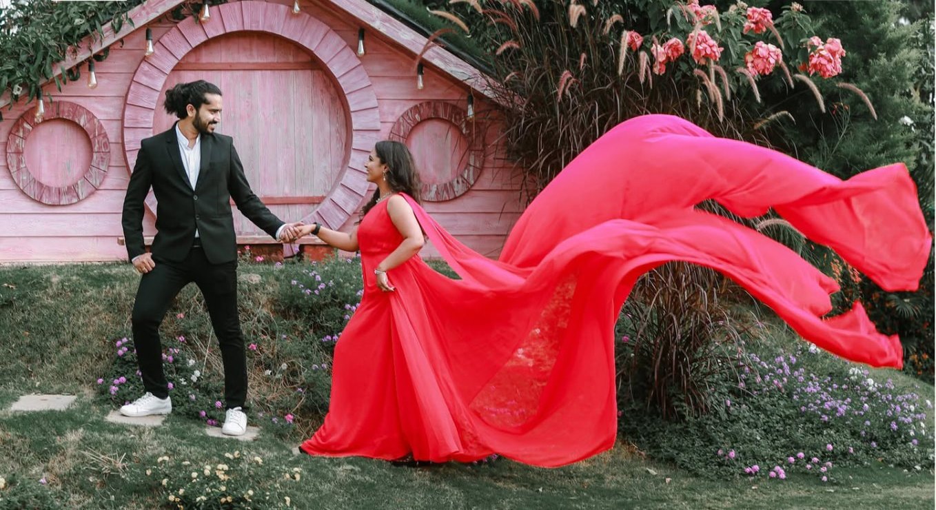 Pre-wedding couple portrait with the bride in a flowing scarlet red gown mid-flight beside a pink wooden cottage and flowering hedges, groom in a black suit — Dar Al Muteena Studio Dubai