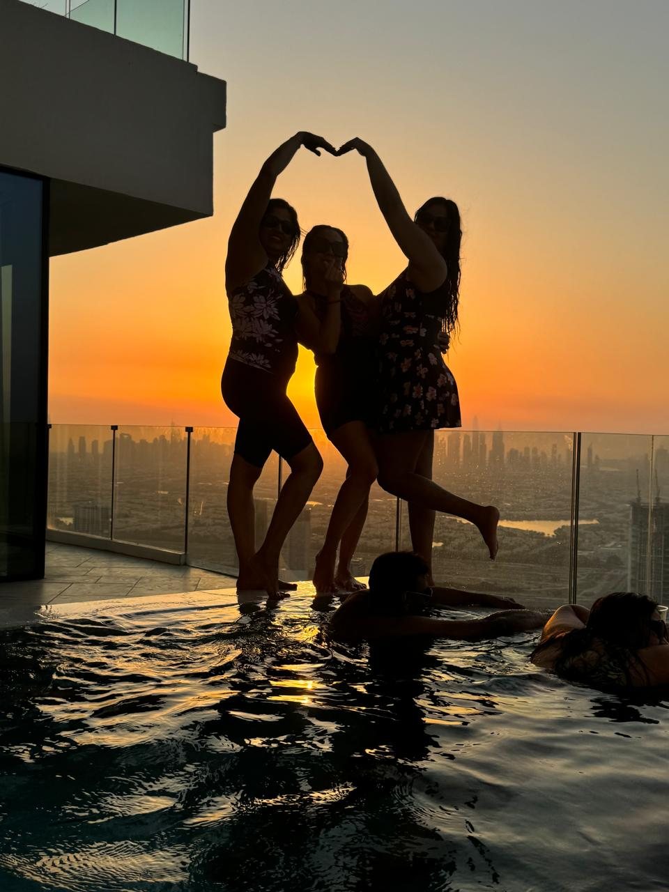 Three friends silhouetted against a Dubai sunset on a rooftop infinity pool, arms raised in a heart shape above the city skyline — lifestyle portrait photography Dubai