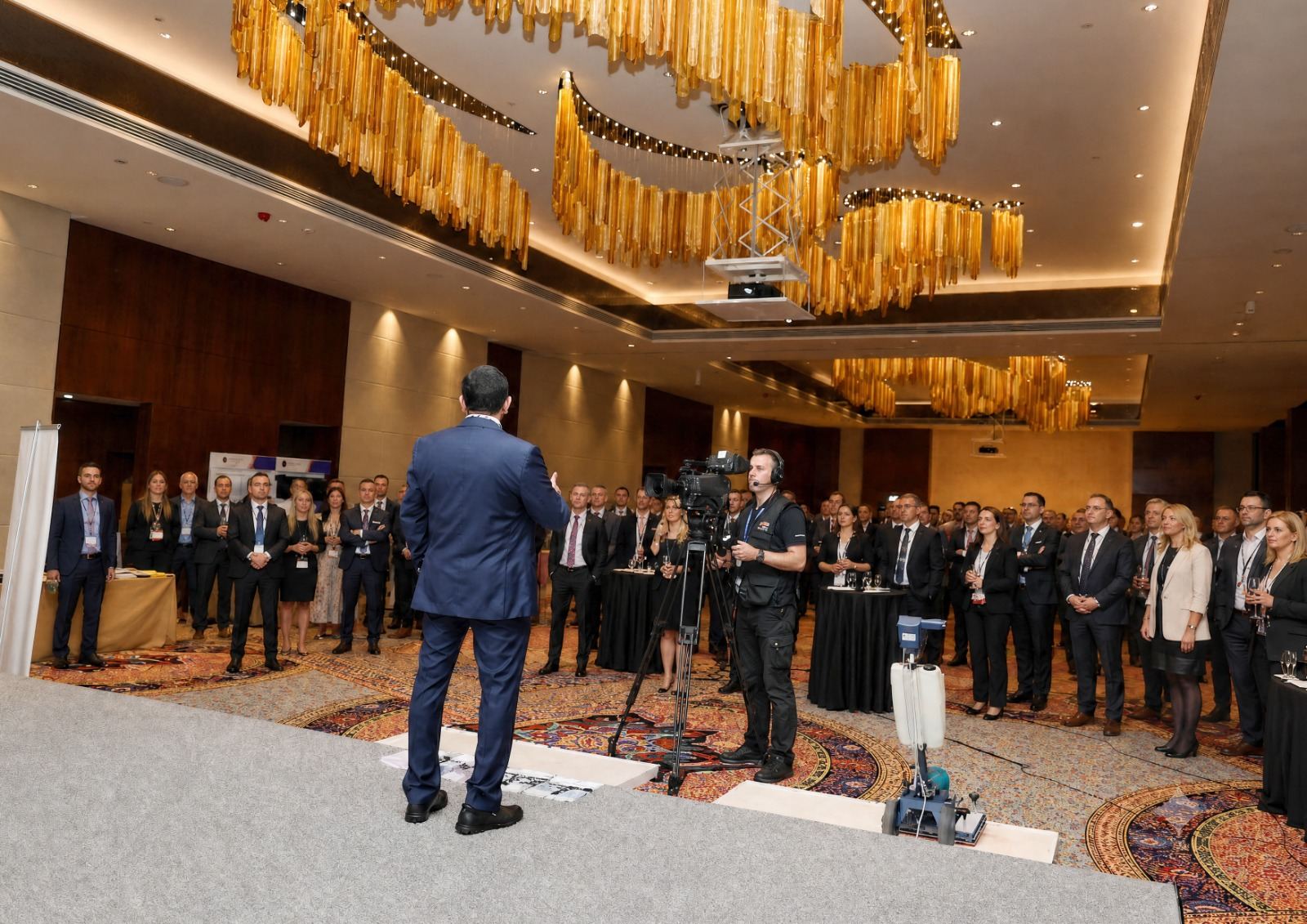 Executive addressing delegates under amber chandeliers at a corporate ballroom conference in Dubai, filmed by a video crew