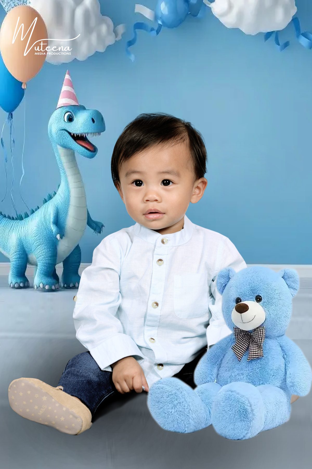 Baby boy in a white shirt seated with a teddy bear and blue balloon dinosaur birthday setup in Deira