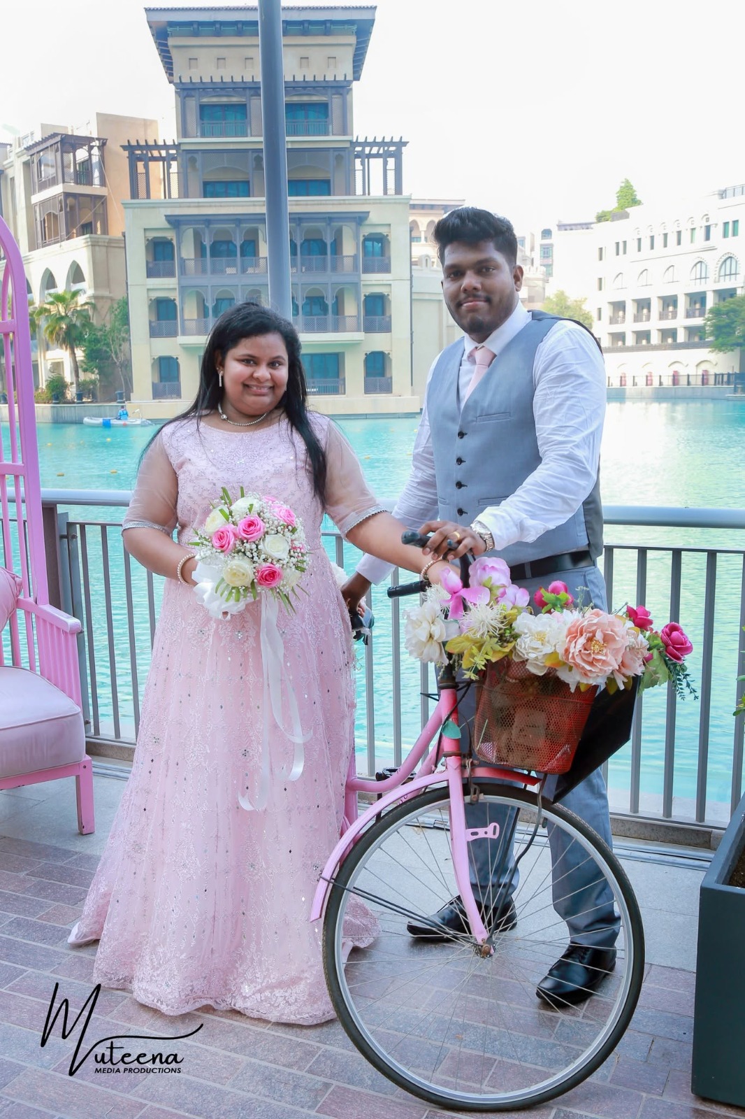 Bride in a blush pink lehenga holding a pastel bouquet beside a pink vintage bicycle and Downtown Dubai fountain — whimsical pre-wedding shoot by Dar Al Muteena Studio