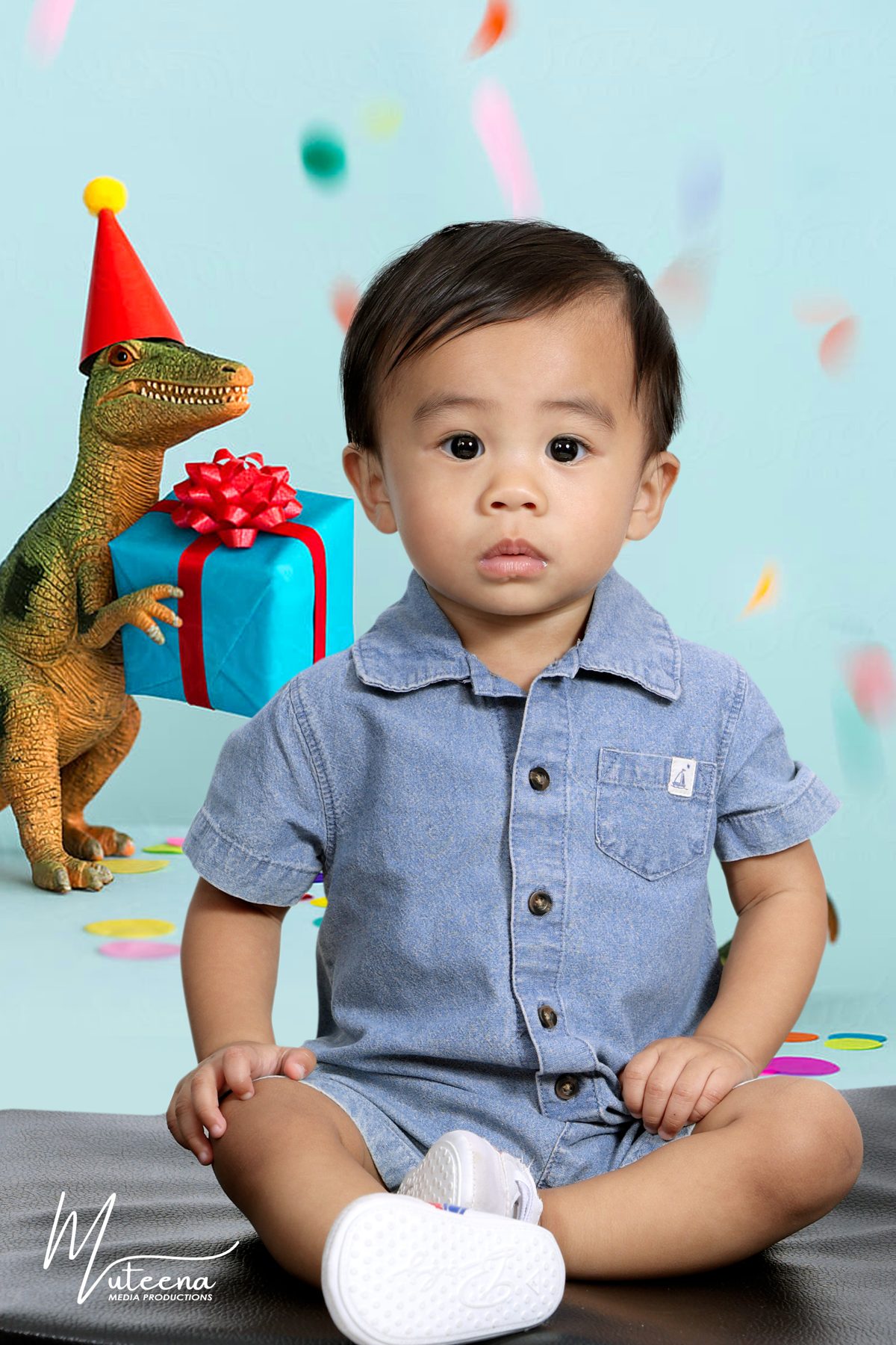 Toddler in denim with a gift box and party dinosaur on a mint confetti backdrop at Dar Al Muteena Studio